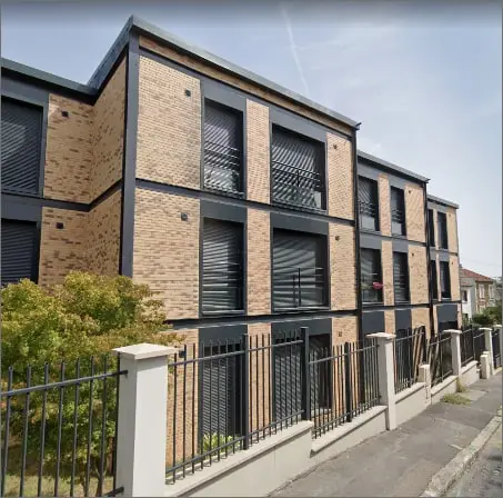 La façade en brique claire avec volets roulants sombres et clôture métallique sur rue — chantier à Montreuil.