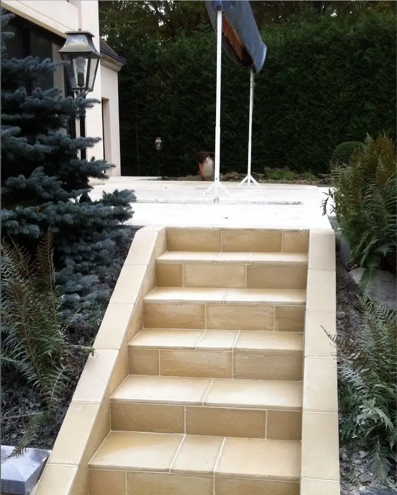 L’escalier extérieur carrelé de couleur beige, bordé de murets clairs et de végétation.